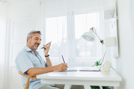 Mature adult man using voice recognition on mobile phone while working on laptop computer at home. Talking on Phone, Speaker Phoneの写真素材