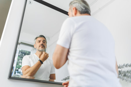 Portrait of handsome Mature adult man touching his beard and looking at his reflection in mirrorの写真素材