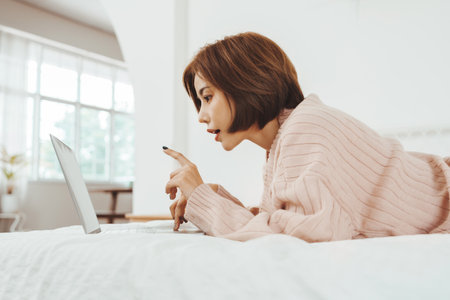 Young Beautiful Asian Korean woman lying on the bed and having online shopping on a laptop, Chatting and happy smiling, Working at homeの写真素材