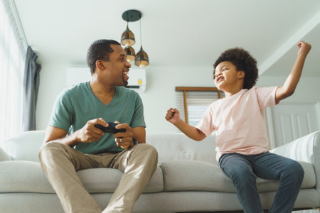 Cheerful Black African American Father and his Little son in Afro hair sitting on sofa using...