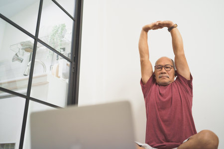 Asian Senior man stretching his arms before yoga exercise online at home. Using Laptop.の写真素材