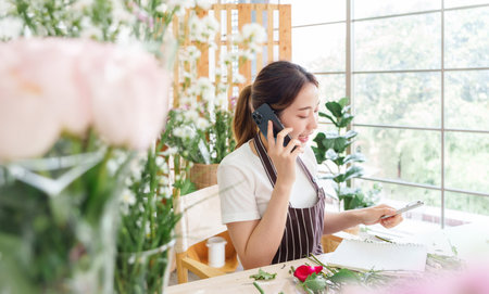 Beautiful Asian florist woman in apron talking on mobile phone  with clients or customers at flower shop, Female receiving order of flowers, Small business, entrepreneur conceptの写真素材