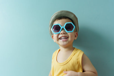 Portrait of Cheerful little Asian boy in yellow sleeveless and blue sunglasses on blue background. Smiling faceの写真素材