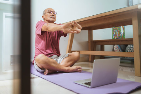 Asian Senior adult man in eyeglasses stretching his arms after home exercise at home. Yoga Online classroom on laptop.の写真素材
