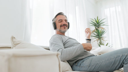 Senior mature adult man sitting on sofa listening to music with headphones and playing an air guitar, Lifestyle concept, Relaxation at home.の写真素材