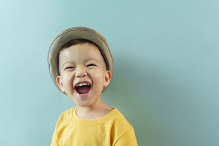 Portrait of Cheerful little Asian boy in yellow shirt and hat laughing standing on isolated blue...