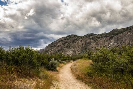 Dirt Trail Winding through the Desert towards the Mountains under a Cloudy Skyの写真素材