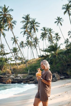 Woman Drink Coconut Juice at Tropical Beach. Happy Lady Enjoy Exotic Fresh Coco Cocktail on Seascape with Palm Rock. Female Tourist Smile. Summer Travel Paradise Landscapeの写真素材