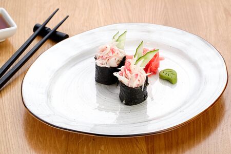 Japanese Sushi Rolls with Crab Meat and Cucumber, Ginger and Wasabi on White Plate. Soy Sauce in Bowl and Eating Sticks on Wooden Table. Healthy Oriental Rice Dish Meal Food Closeup Photoの写真素材