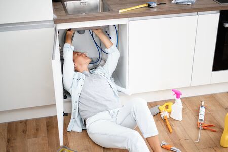 Woman fixing kitchen sink. Housewife working under kitchen sink, tools in foregroundの写真素材
