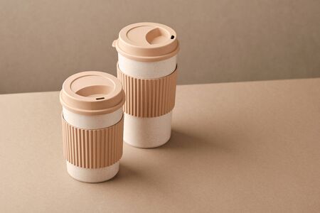 Two Eco Coffee Cup Isolated on Beige Background Closeup Copy Space Photo. Stylish Paper Container Different Size for Morning Hot Energy Drink. Disposable Eco-friendly Cardboard Mugs for Beveragesの写真素材