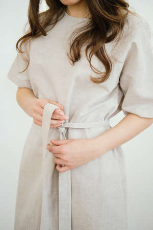 Woman with long dark hair ties a belt on a light gray linen dress. Front view.の写真素材