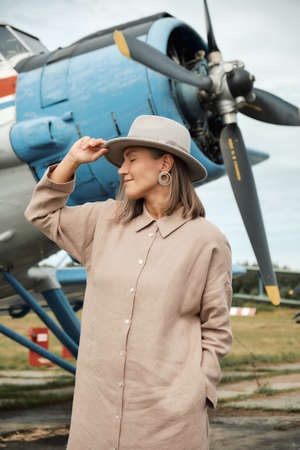 Beautiful girl in a beige linen shirt dress and hat stands against the background of an old plane, closed her eyes and dreams of flying away on a trip. Front view.の写真素材