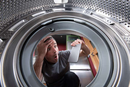 Worried caucasian man taking out a phone from the drum of the washing machine. Interior view.の写真素材