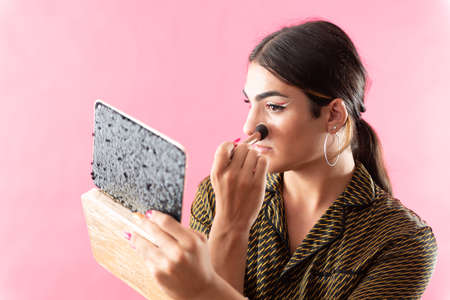 Studio portrait with pink background of a young man putting on makeup while looking in the mirror.の写真素材