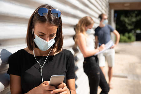A young woman wearing a mask and chatting on her cell phone in a queue during Coronavirus or Covid-19 pandemic.の写真素材