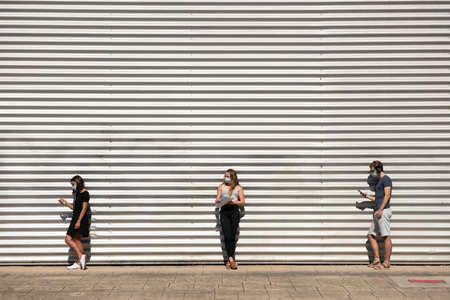 Three young students waiting in line on a queue wearing face masks and keeping social distance. Metallic wall background with copy spaceの写真素材