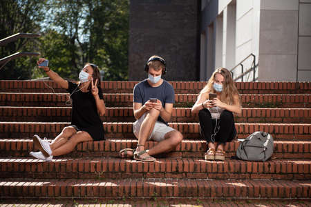 Social distancing concept. Three college students sitting on the stairs and using their smartphones wearing a face mask and keeping social distance during coronavirus pandemic.の写真素材