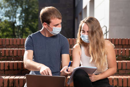 Back to the university during coronavirus pandemic. Two college students with masks pointing to a laptop while sitting on the stairs.の写真素材