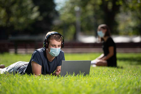 Back to college during Coronavirus or Covid-19 pandemic. A handsome young student using his laptop on the ground of a park while wearing a face maskの写真素材