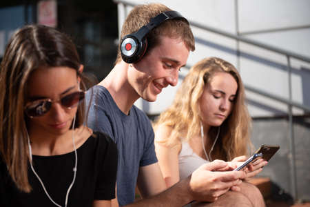 Digital technology addiction concept. Caucasian blond young boy whearing headphones and smiling while chatting surrounded by two girls.の写真素材