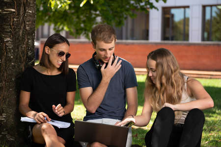 Three college students working together on a laptop under a tree in the park of the campus.の写真素材