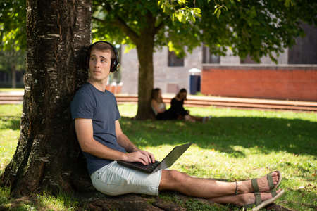A blond young male student working on his laptop sitting under a tree in the park.の写真素材