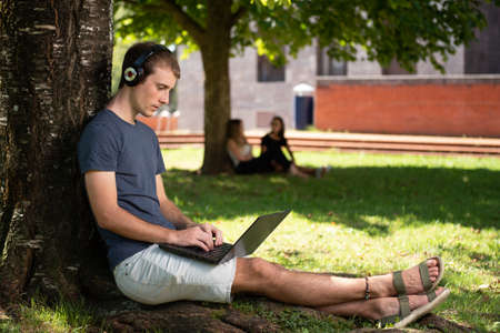 A blond young male student working on his laptop sitting under a tree in the park.の写真素材