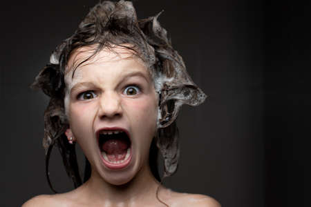 Closeup portrait of a horrified little girl in the shower with soap foam in the hair. Child health care and hygiene at bath time concept.の写真素材