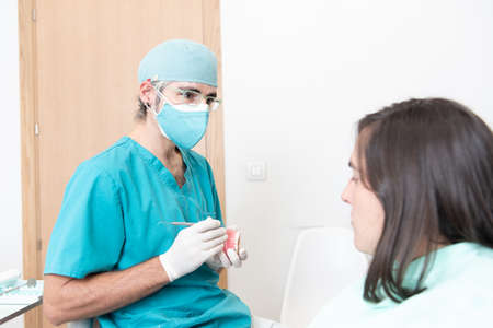 Male dentist explaining the situation to a patient using a false dentition during Coronavirus pandemic with double face mask.の写真素材
