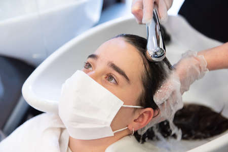 Everyday life during Coronavirus pandemic concept. Close up view of a hairstylist washing the hair to a teenager patient wearing a face mask.の写真素材