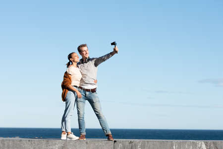 Social media content creation concept. A young couple taking themselves a selfie with a compact camera in a sunny day at the seaside.の写真素材