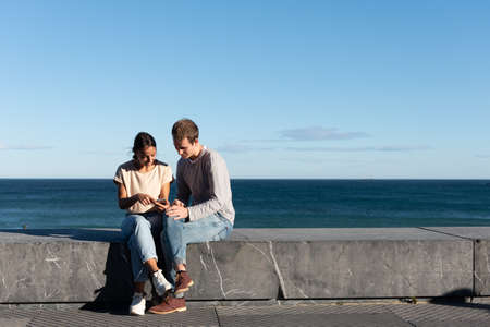 Young couple sitting on a stone seat and checking their smartphone at the seaside in a sunny dayの写真素材