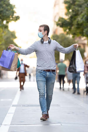 Shopping during Coronavirus concept. A handsome boy walking through a pedestrian street holding his shopping bags and wearing a face mask.の写真素材
