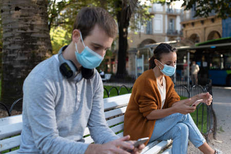 Young man and woman sitting on a bench checking their phones while keeping social distance during Coronavirus pandemic. Couple wearing face masksの写真素材
