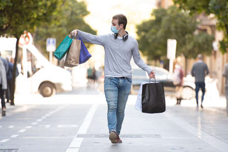 Shopping during Coronavirus concept. A handsome man walking through a pedestrian street holding his shopping bags and wearing a face mask.の写真素材