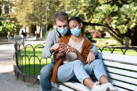 Young couple in romantic position sitting on a bench. Lovers checking a phone wearing face masks during Coronavirus pandemic.の写真素材