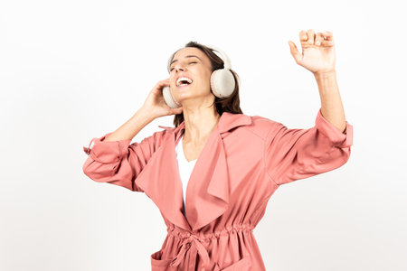 Happy latin woman wearing headphones and a raincoat dancing over white background.の写真素材