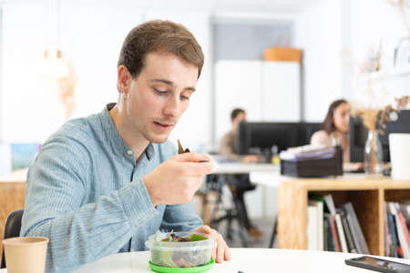 Healthy food in the workplace concept. Handsome worker having a lunch break and eating a salad in the office.の写真素材