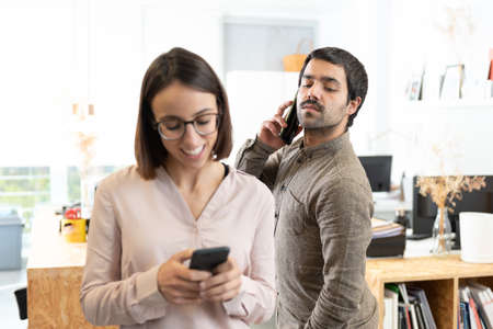 Hispanic man with mustache spying the phone of his female coworker while she is chatting.