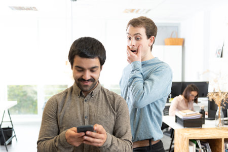 Surprised young man spying a coworker while he is looking at his cell phone at the office.の写真素材