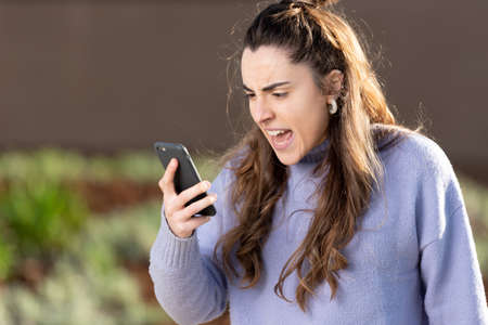 Angry young caucasian woman looking at her phone outdoors. Social media and mental health conceptの写真素材