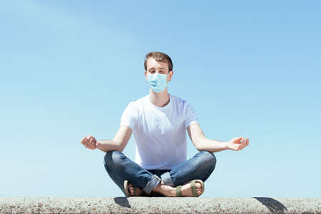 Meditation, yoga and mental health during Coronavirus concept. Handsome man sitting in lotus pose outdoors and wearing a protective mask.の写真素材