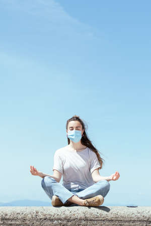 Meditation, yoga and mindfulness during Coronavirus concept. Young woman sitting in lotus pose and wearing a protective mask.の写真素材