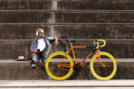 Happy hispanic man with blond hair sitting on stands and listening to music beside a fixie colorful...