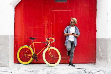 Hispanic man against an old red door holding his phone beside his vintage bicycle. Sustainable tourism and mobility conceptの写真素材
