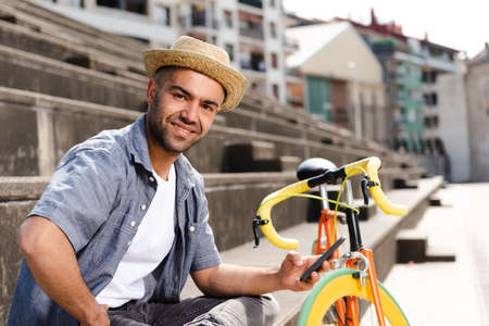 Smiling hispanic man holding his phone and looking to the camera beside a vintage bicycle.の写真素材