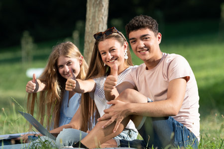 Successful happy students gesturing thumbs up sitting outdoors on the ground of a park.の写真素材
