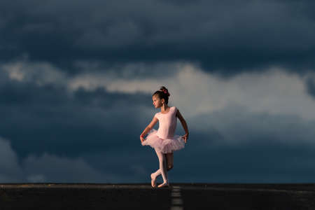 Little ballet dancer girl wearing a pink tutu in the horizon of the road at the golden hour. Dramatic stormy sky with copy space in the backgroundの写真素材