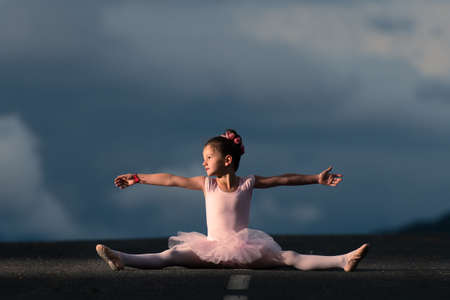 Little ballet dancer girl wearing a pink tutu doing a split on floor on the road. Dramatic sky with copy space in the backgroundの写真素材